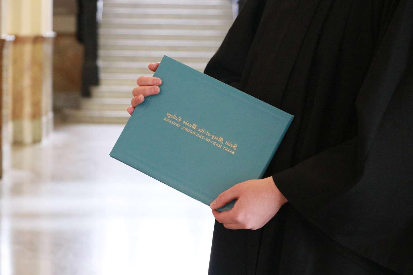 The hands of someone in a graduation gown holding a degree jacket that says Saint Mary-of-the-Woods College.